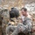 A U.S. Army soldier in MultiCam/OCP camouflage uniform and helmet is providing medical assistance to a seated casualty in ACU (Army Combat Uniform) pattern clothing. The casualty appears shirtless from the waist up with what appears to be simulated wound makeup on his leg, suggesting this is a combat medical training exercise. The setting is an outdoor field with dried vegetation and patches of snow or ice visible in the background.