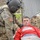 A U.S. Army Airborne soldier in full combat gear and flight helmet is attending to a simulated casualty during what appears to be a training exercise. The 'casualty' is a young male in civilian clothing lying on the ground near a chain-link fence, with simulated blood (moulage) applied to his face and clothing. The scene is consistent with a combat casualty care or MEDEVAC training scenario.