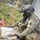 A military training exercise depicting a combat casualty care scenario. A soldier in full tactical gear and helmet is attending to a simulated casualty wearing moulage (theatrical makeup simulating wounds). At least two 'casualties' are visible with simulated facial injuries, indicating a mass casualty training drill.