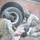 Two soldiers in U.S. Army combat uniforms and tactical helmets are providing medical assistance to a downed civilian casualty near a deteriorated red vehicle. The scene appears to be a military training exercise involving combat casualty care (TCCC). The soldiers are equipped with tactical gear including backpacks and body armor.