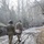 Two soldiers in military combat uniforms and helmets are carrying a casualty on a stretcher down a muddy, partially snow-covered path flanked by chain-link fencing and bare trees. The scene appears to be a military training exercise, with additional personnel and military vehicles visible in the background. Hazy atmospheric conditions, possibly from smoke, reduce visibility in the mid-ground.