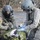 Two U.S. Army soldiers in full combat gear and flight helmets are providing medical treatment to a casualty lying on a thermal/emergency blanket on the ground. Both soldiers are wearing American flag patches and one has an Airborne tab visible on their left shoulder. The scene appears to be a field medical training exercise or real casualty care operation in an outdoor environment.