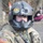 A U.S. military personnel wearing a flight/aviation helmet equipped with a GoPro Hero4 camera and a Teenage Mutant Ninja Turtles 'Cowabunga' patch. The individual is dressed in MultiCam (OCP) camouflage uniform with a U.S. flag patch on the left sleeve and tactical gear. In the blurred background, at least one civilian observer can be seen crouching near a wooded area.