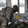 A U.S. Army soldier in full combat gear and flight helmet is leaning over what appears to be a patient or casualty wrapped in a thermal/emergency blanket inside a military helicopter. The soldier appears to be performing a medical assessment or treatment, suggesting this is a MEDEVAC (medical evacuation) operation or training exercise. The helicopter interior and open door reveal a grassy field in the background.