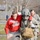 The image depicts what appears to be a military medical training exercise or disaster response drill. Two military personnel in Army Combat Uniforms (ACU) — one wearing a flight helmet — are assisting a simulated casualty wearing a red hoodie with visible mock injuries (simulated blood/moulage). Additional personnel and a fenced compound are visible in the background.