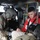 A U.S. Army medic wearing a flight helmet and combat uniform is providing medical treatment to a casualty inside what appears to be a military helicopter, likely a UH-60 Black Hawk. The casualty, wearing a red sweatshirt, has simulated wounds on his face and leg, suggesting this is a medical training exercise. The medic is attending to the patient's lower extremity while the patient is secured in a seat.