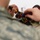 A person wearing US Air Force ABU (Airman Battle Uniform) camouflage is assembling or repairing a small wheeled robot or electronic vehicle. They are using a screwdriver to work on the device, which contains visible wiring, circuit boards, and rubber wheels. A printed document or schematic is visible in the background on the work surface.