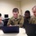 Four U.S. Air Force personnel in military uniforms are gathered around laptops in what appears to be a training or computer lab environment. Three individuals in OCP (Operational Camouflage Pattern) uniforms and one in ABU (Airman Battle Uniform) are collaborating on a task displayed on a laptop screen. Name tapes visible include 'BARGER' and partially visible others on the right side of the group.