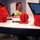 Three red 3D-printed objects are displayed on a white table in what appears to be a makerspace or design studio. The objects include a detailed wolf head bust, a geometric sphere with interlocking curved panels on a small base, and what appears to be a pig or animal face cup/container. A person in a yellow-green shirt is seated in the background working at a computer monitor.