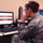 A military personnel member in camouflage uniform is seated at a workstation reviewing data on a computer monitor. The screen displays what appears to be a spreadsheet or database application with red-highlighted rows, suggesting alerts or flagged entries. The setting appears to be a military operations or cyber operations room.