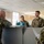 Four U.S. Army soldiers in OCP (Operational Camouflage Pattern) uniforms are engaged in a conversation in what appears to be an office or administrative setting. Two soldiers stand behind a cubicle partition while two others face them from the opposite side. The interaction appears informal and positive, with visible smiling among the personnel.