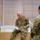 Three U.S. Army soldiers in Operational Camouflage Pattern (OCP) uniforms are gathered in what appears to be an indoor facility, reviewing documents. A senior soldier, seated on a ledge, is reading from a booklet while two other soldiers stand nearby holding additional papers. The setting suggests a briefing, planning session, or administrative review.