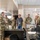 A group of U.S. Army personnel in OCP (Operational Camouflage Pattern) uniforms are gathered in what appears to be a military operations or command center. One soldier appears to be briefing or presenting information to the group, which also includes one civilian male wearing a badge/lanyard. A large map is mounted on the wall in the background.
