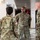 Four U.S. Army soldiers in Operational Camouflage Pattern (OCP) uniforms are engaged in a conversation in an indoor hallway of a military facility. The soldier facing the camera has a name tape reading 'FREDERICK' and a 'U.S. ARMY' tape visible on his uniform. The setting appears to be a military building with standard institutional features including an EXIT sign, fire safety signage, and bulletin boards.