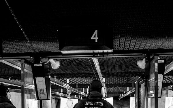 A United States Coast Guard member stands at lane number 4 of an indoor shooting range, facing downrange toward paper silhouette targets. The image is captured in black and white, showing the shooter from behind in full tactical black attire. The facility appears to be a professional multi-lane indoor firearms training range.