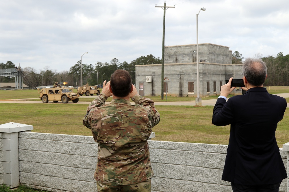 Two observers — one in military camouflage uniform and one in a civilian business suit — stand behind a low concrete block wall watching and photographing a military vehicle demonstration. A tan HMMWV (Humvee) with a mounted weapon system is visible in the background on a training range. The setting appears to be a military installation with a concrete block building and utility poles in the background.
