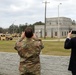 Two observers — one in military camouflage uniform and one in a civilian business suit — stand behind a low concrete block wall watching and photographing a military vehicle demonstration. A tan HMMWV (Humvee) with a mounted weapon system is visible in the background on a training range. The setting appears to be a military installation with a concrete block building and utility poles in the background.