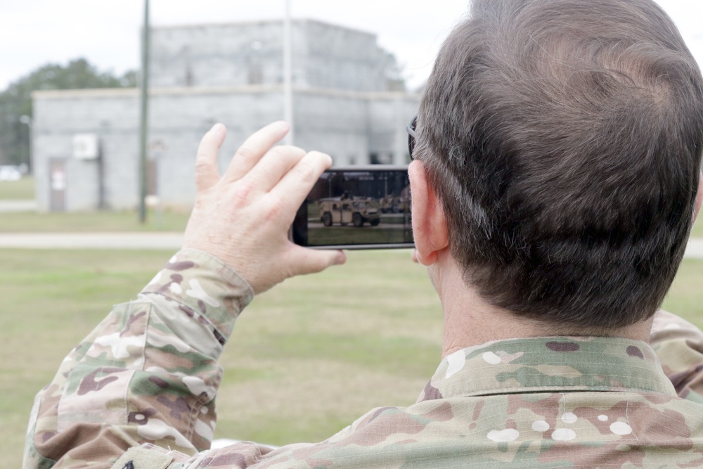 A military personnel member wearing MultiCam/OCP camouflage uniform is photographing a military vehicle using a smartphone. The photo is taken from behind the subject, showing both the person and the vehicle captured on the phone screen. The setting appears to be a military installation or training area with structures visible in the background.