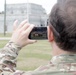 A military personnel member wearing MultiCam/OCP camouflage uniform is photographing a military vehicle using a smartphone. The photo is taken from behind the subject, showing both the person and the vehicle captured on the phone screen. The setting appears to be a military installation or training area with structures visible in the background.