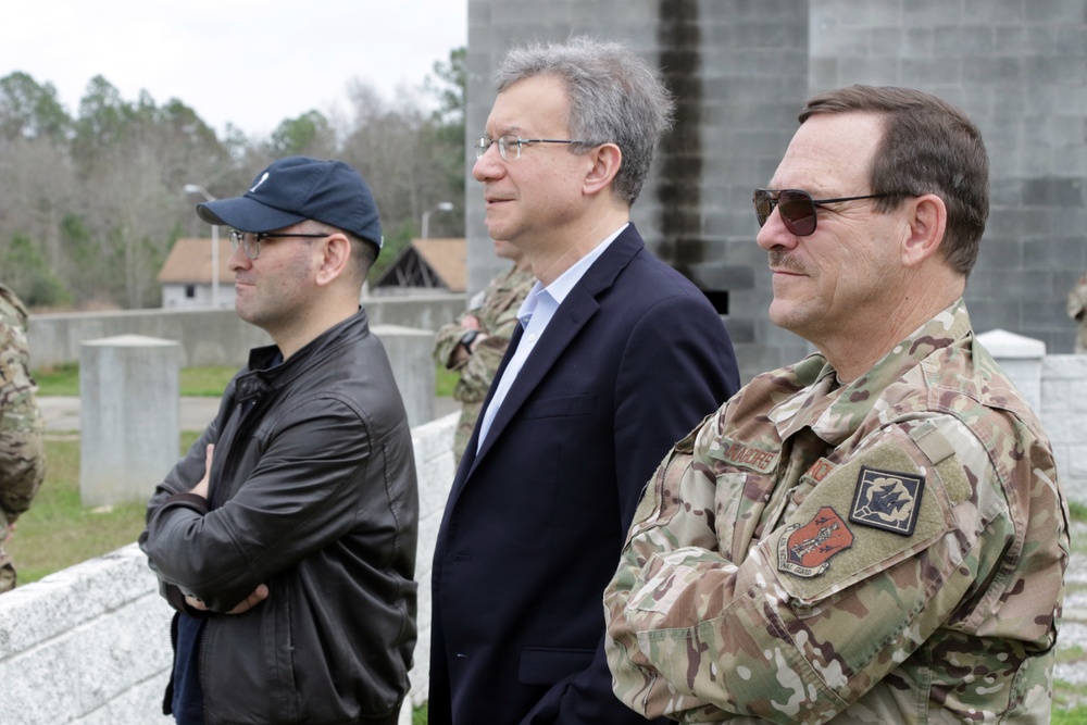 Three men stand together outdoors at what appears to be a military installation or training facility. Two individuals are in civilian attire while one is wearing U.S. military camouflage (OCP/MultiCam) uniform with unit patches visible on the sleeve. They appear to be observing something off-camera, possibly a demonstration or briefing.