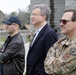 Three men stand together outdoors at what appears to be a military installation or training facility. Two individuals are in civilian attire while one is wearing U.S. military camouflage (OCP/MultiCam) uniform with unit patches visible on the sleeve. They appear to be observing something off-camera, possibly a demonstration or briefing.