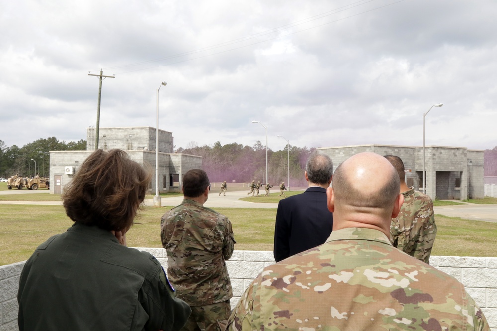 A group of military personnel and at least one civilian are observing a tactical training exercise at what appears to be a military training facility. Soldiers in the background are conducting a live demonstration involving purple smoke grenades near concrete training structures. The observers are positioned behind a low concrete barrier watching the exercise unfold.