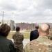 A group of military personnel and at least one civilian are observing a tactical training exercise at what appears to be a military training facility. Soldiers in the background are conducting a live demonstration involving purple smoke grenades near concrete training structures. The observers are positioned behind a low concrete barrier watching the exercise unfold.