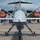 A front-facing view of a military unmanned aerial vehicle (UAV), likely an MQ-9 Reaper, displayed on a tarmac at what appears to be an air show or static display event. Behind the drone, a large US Air Force transport aircraft (likely a C-17 Globemaster III) is visible with red engine covers installed. The setting is an outdoor aviation exhibition with security barriers and spectators visible.