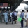 The image shows a military airshow or aviation exhibition where US Air Force personnel in olive flight suits are briefing a group of civilian and military visitors near a large military transport aircraft. An American flag is displayed inside the open cargo bay of the aircraft, which has white chairs set up inside, suggesting a formal display or reception area. A metal crowd-control barrier separates the crew from the visitors.