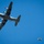 A USAF C-130 Hercules military transport aircraft is photographed from below against a clear blue sky, actively conducting an airdrop operation. Cargo packages with parachutes are visible having been released from the aircraft's rear ramp. The image captures the moment of deployment with at least two cargo bundles visible in freefall or with parachutes deploying.