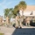 A group of military personnel in camouflage uniforms are gathered on a paved area outside a building, appearing to prepare for a parachute or airborne operation. Multiple soldiers are donning or adjusting parachute rigs and equipment, with gear bags scattered across the ground. The setting appears to be a military installation, likely in a warm climate based on the palm trees and vegetation visible in the background.