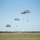 A military airborne operation is underway over a large open grassy field, with multiple paratroopers descending under olive-drab/gray round parachutes. At least four parachutes are visible at varying altitudes, with some paratroopers nearly at ground level. A tree line and urban buildings are visible in the background horizon.