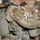 A U.S. Army Airborne soldier is shown from behind/side wearing Operational Camouflage Pattern (OCP) uniform with a parachute rig and harness system. Visible patches include an American flag, an 'Airborne' tab, and a unit shield patch on the left shoulder. The soldier appears to be in a pre-jump staging or inspection area with other personnel visible in the blurred background.