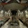 The interior of a large military cargo aircraft, likely a C-17 Globemaster III, is filled with rows of soldiers in full combat gear seated along both sides of the fuselage. The troops appear to be equipped with parachute rigs and tactical equipment, suggesting a airborne operation or jump mission. The wide-angle perspective reveals the full length of the cargo bay with soldiers packed tightly on both port and starboard seating.
