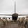 A large group of military paratroopers in full combat gear and parachute equipment are lined up in two columns on a tarmac, boarding a military transport aircraft through its rear cargo ramp. The soldiers are wearing MultiCam/OCP uniforms with helmets and are equipped with green parachute packs and yellow static lines. The scene appears to be a pre-jump load operation at a military airfield under overcast skies.