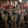 A group of military personnel in full combat gear are seated inside what appears to be a military transport aircraft, likely a C-130 or similar cargo plane. The soldiers are wearing camouflage uniforms, helmets, and parachute harnesses, suggesting they are preparing for or returning from an airborne operation. The atmosphere appears relatively relaxed, with some personnel conversing and smiling.