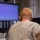 CBP Support to Super Bowl LIV A person with short blonde/reddish hair and glasses is seated at a workstation viewing a large wall-mounted monitor displaying what appears to be aerial surveillance or drone footage with green and red tactical overlays. A second LG monitor is visible to the right showing what appears to be an email client. The setting appears to be a military or law enforcement operations center.