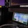 CBP Support to Super Bowl LIV A ruggedized military-grade tablet in an olive/green protective case is displaying a tactical mapping application, likely ATAK (Android Tactical Assault Kit) or similar military situational awareness software. The device is positioned on a dark surface inside what appears to be a military vehicle or command station, with larger display screens visible in the background. A flexible metal conduit cable is connected to the device.