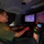 CBP Support to Super Bowl LIV A U.S. Border Patrol agent is seated inside a surveillance vehicle, operating multiple monitoring screens and control equipment. The agent is wearing a green Border Patrol uniform with a visible shoulder patch and is actively working at a workstation with dual monitors displaying thermal/infrared camera feeds. The setup appears to be a mobile command or surveillance unit used for border monitoring operations.