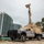 CBP Support to Super Bowl LIV A military or law enforcement surveillance vehicle equipped with a tall extendable mast-mounted sensor/camera system is parked on sandy ground near a coastal urban area. The vehicle appears to be a modified Ford F-450/550 heavy-duty truck with tan/desert-colored equipment mounted in the bed. A white government SUV, likely U.S. Customs and Border Protection, is visible in the background alongside modern high-rise buildings.