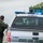 CBP Support to Super Bowl LIV A U.S. Border Patrol police officer stands beside a Ford F-150 pickup truck marked 'State Law Enforcement' with active blue emergency lights. The officer appears to be conducting a traffic stop or checkpoint inspection, facing toward the vehicle's driver-side window. In the background, a large truck loaded with what appears to be agricultural produce (corn or similar crop) is visible.