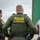 CBP Support to Super Bowl LIV Two U.S. Border Patrol officers in olive green tactical uniforms with 'POLICE - U.S. BORDER PATROL' markings on their vests are standing near a large green truck or trailer. A civilian male in a gray t-shirt and black cap is visible to the right, appearing to interact with or be in proximity to the officers. The scene appears to take place outdoors under overcast skies, suggesting a field operation or vehicle stop.