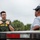 CBP Support to Super Bowl LIV A U.S. Border Patrol Federal Agent in green tactical uniform and body armor is engaged in a conversation with a civilian near what appears to be a vehicle or trailer. The agent is gesturing toward his chest/vest area, possibly explaining something or pointing to his credentials. The setting appears to be an outdoor location with overcast skies and vegetation in the background.