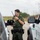 CBP Support to Super Bowl LIV A U.S. Border Patrol Federal Agent stands between two government vehicles, speaking into a handheld radio while holding what appears to be a tablet or phone. The agent is wearing full tactical gear including a green uniform, body armor vest with 'Border Patrol Federal Agent' markings, and a duty belt. The setting appears to be an outdoor staging area near a field with power transmission towers visible in the background.