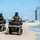 CBP Support to Super Bowl LIV Two law enforcement officers wearing green uniforms and tactical gear are patrolling a sandy beach on olive-green ATVs (all-terrain vehicles). Both officers are wearing full-face helmets and body armor vests with visible agency patches. A modern urban skyline with high-rise buildings is visible in the background along the coastline.