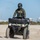 CBP Support to Super Bowl LIV A U.S. Border Patrol Federal Agent is riding a green Can-Am ATV on a sandy beach. The agent is wearing full tactical gear including a helmet, body armor vest with visible 'Border Patrol Federal Agent' patch, and a sidearm. Buildings and vegetation are visible in the background, suggesting a coastal urban or semi-urban environment.