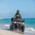 CBP Support to Super Bowl LIV A U.S. Border Patrol agent is riding a green/olive-colored ATV along a sandy beach shoreline with turquoise ocean water visible in the background. The agent is wearing full tactical gear including a black helmet, olive uniform, and a tactical vest with visible Border Patrol insignia. Tall residential or hotel buildings are visible along the coastline in the left background.