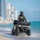 CBP Support to Super Bowl LIV A law enforcement officer wearing a tactical vest with visible 'Border Patrol' insignia is riding a green ATV along the shoreline of a beach. The officer is fully equipped with a helmet, gloves, and body armor, patrolling through shallow surf. A skyline of high-rise buildings is visible in the background, consistent with a South Florida coastal urban environment.