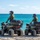 CBP Support to Super Bowl LIV Two law enforcement officers wearing tactical vests labeled 'POLICE' and 'U.S. BORDER PATROL' are seated on olive-green ATVs on a sandy beach facing the ocean. Both officers are wearing helmets and appear to be conducting a coastal patrol. A vessel is faintly visible on the horizon in the background.