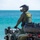 CBP Support to Super Bowl LIV A U.S. Border Patrol police officer is seated on a Can-Am ATV on what appears to be a coastal beach location, looking out toward a turquoise body of water. The officer is in full tactical gear including a black helmet, green uniform, and a tactical vest labeled 'POLICE U.S. BORDER PATROL'. A vessel is faintly visible on the horizon in the background.